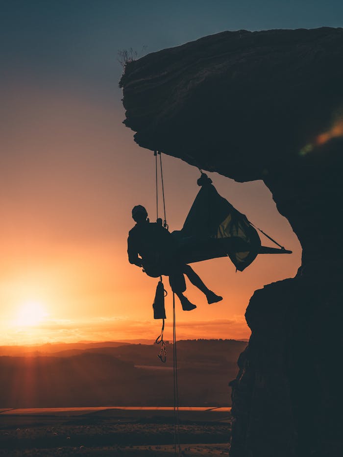 Silhouette of a rock climber hanging from a cliff edge during a colorful sunset, embodying adventure and exploration.